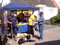 Der Infostand mit einigen Betreuern. Auf dem Tisch liegen Broschüren und Hilfsmittel aus. Im Vordergrund macht es sich ein Führhund bequem. Technischer Hinweis: In der Bildmitte befindet sich ein Hotspot. Wenn Sie darauf klicken, wird das bild vergrößert dargestellt. 